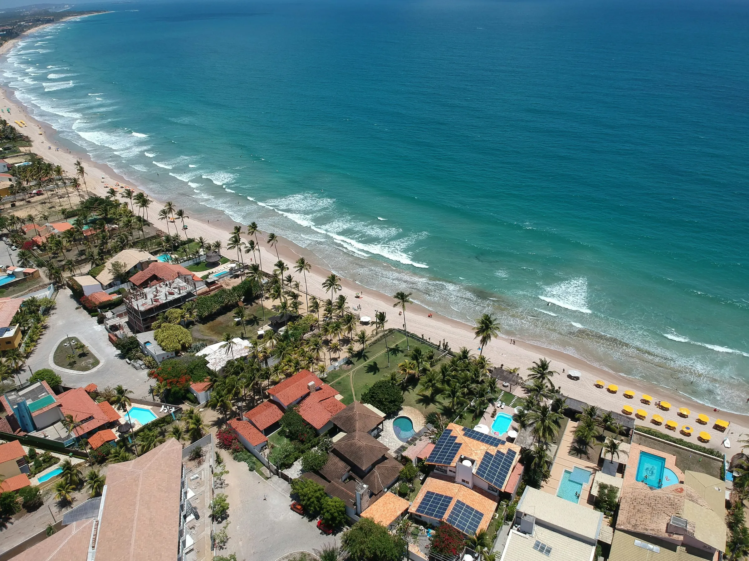 Vista aérea de praia em Porto de Galinhas, com mar azul-turquesa, faixa de areia, coqueiros e casas com piscina