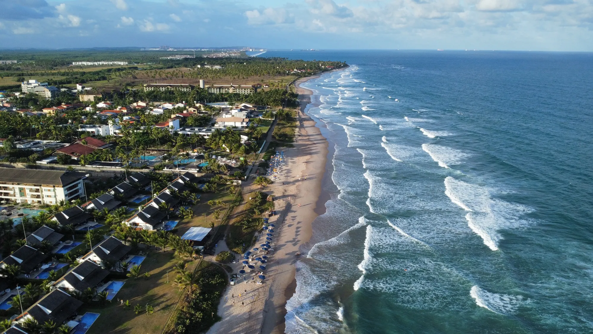Vista aérea da orla de Porto de Galinhas, com ondas quebrando na faixa de areia e coqueiros entre as casas e pousadas