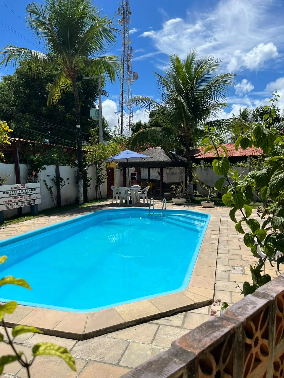 Piscina ao ar livre cercada por coqueiros e jardim, com mesas sob ombrelone